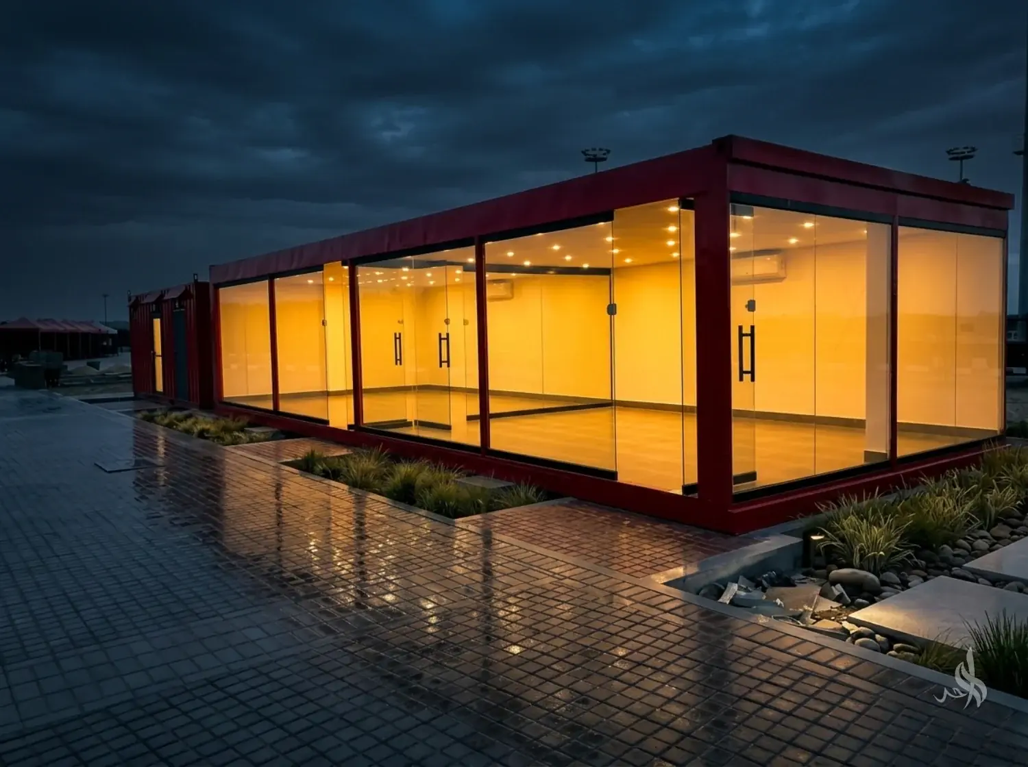 A red-framed container building with full-height glazing, lit warm inside, on wet paving at dusk.